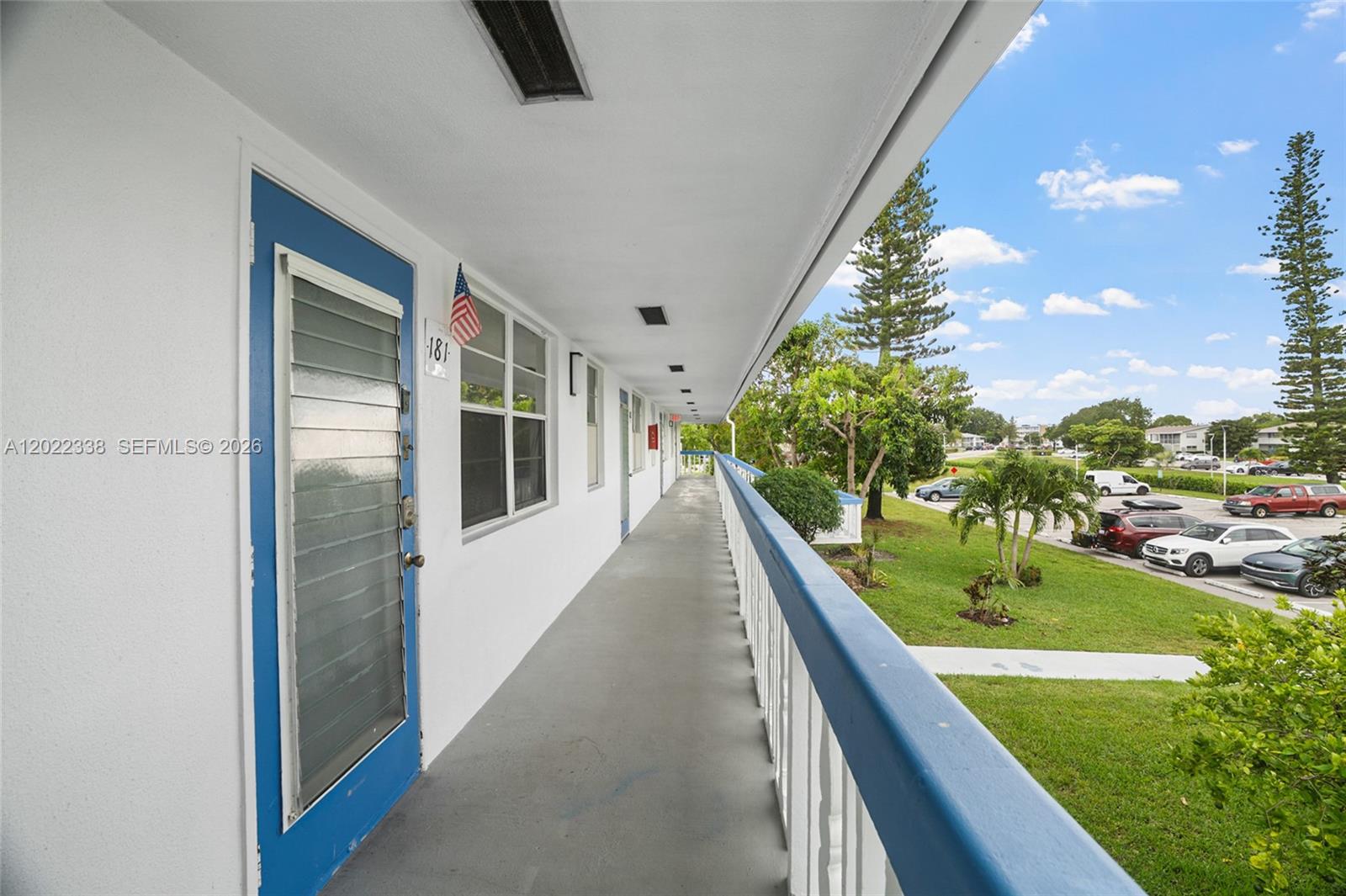 An outdoor walkway with blue doors and railings overlooks a parking lot and green space — 181 Lyndhurst L # 181, Deerfield Beach FL 33442, DEERFIELD BEACH, FL