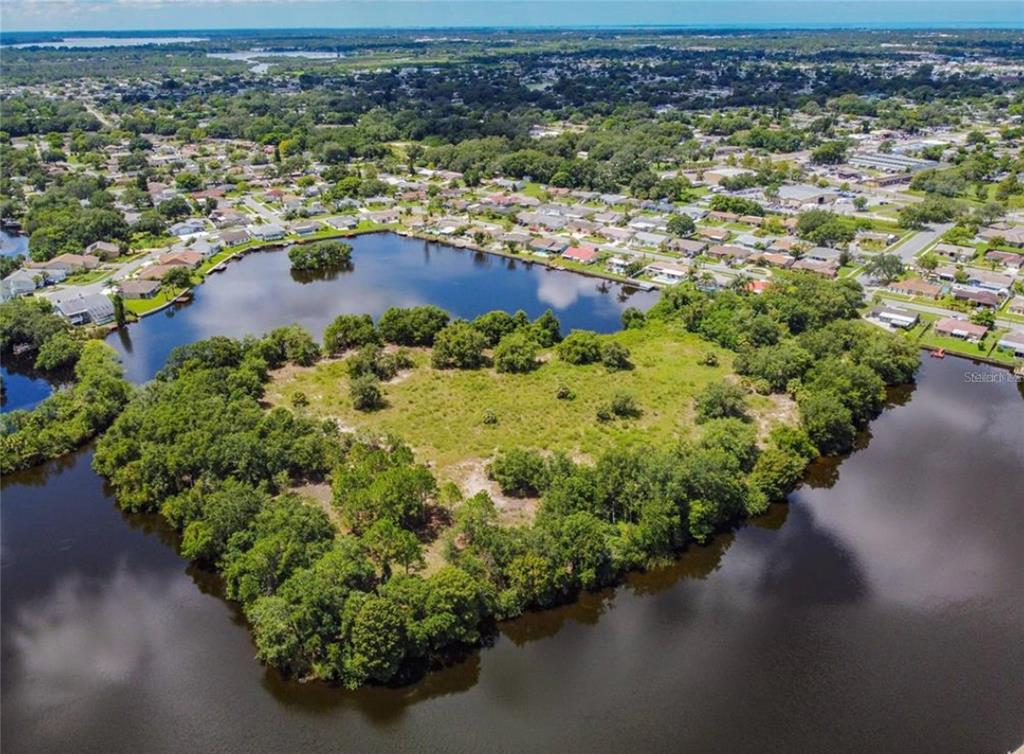 An aerial view shows a grassy, tree-covered peninsula extending into a large lake near houses — 0 BERRY HILL ROAD, HOLIDAY, FL