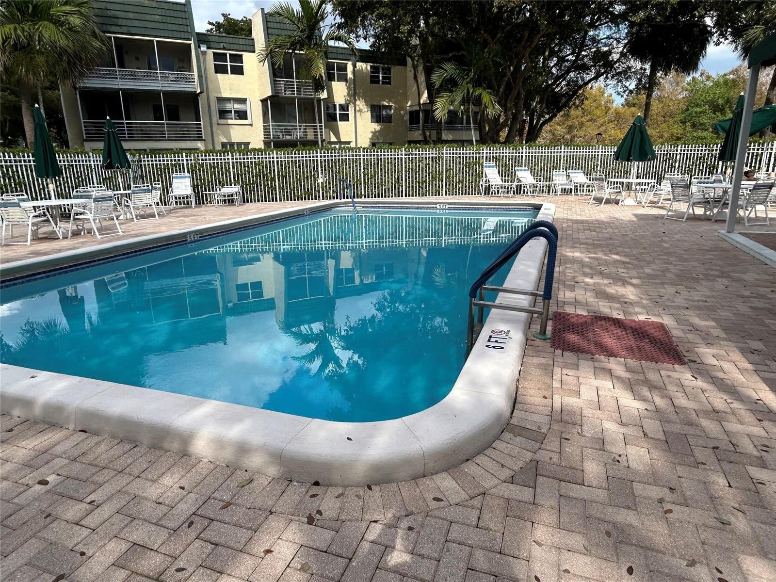 A rectangular swimming pool with a ladder and patio furniture sits before an apartment building — 9080 Lime Bay Boulevard # 305, Tamarac FL 33312, TAMARAC, FL