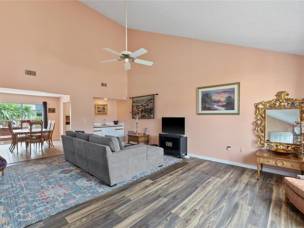 A living room with vaulted ceilings, wood-look flooring, a gray sectional, and a dining area — 3910 77TH PLACE E, SARASOTA, FL