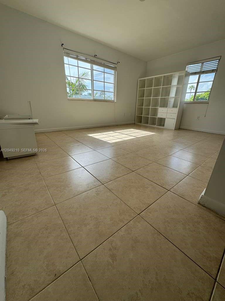 An empty room features beige tile flooring, white walls, two windows, and a white shelving unit — 7130 Rue Versailles # 6, Miami Beach FL 33141, MIAMI BEACH, FL