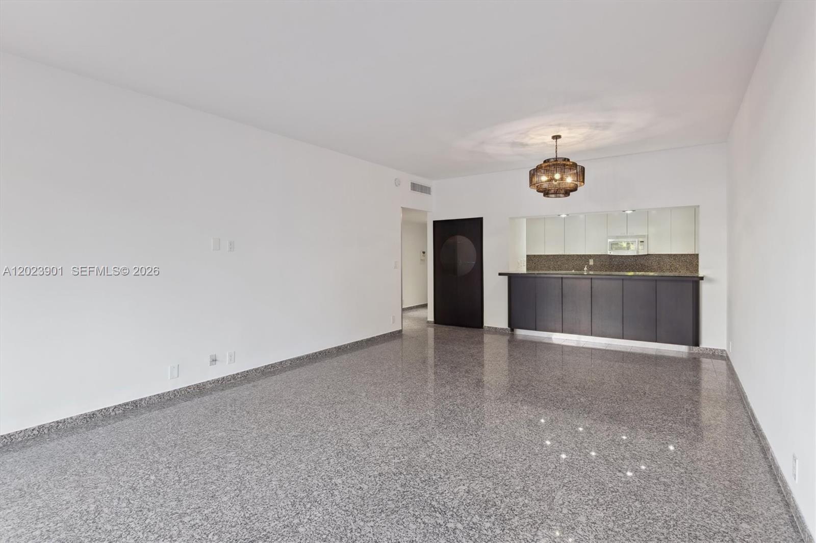 An empty room features gray speckled flooring, white walls, and a dark kitchen bar area — 151 Crandon Blvd # 120, Key Biscayne FL 33149, KEY BISCAYNE, FL