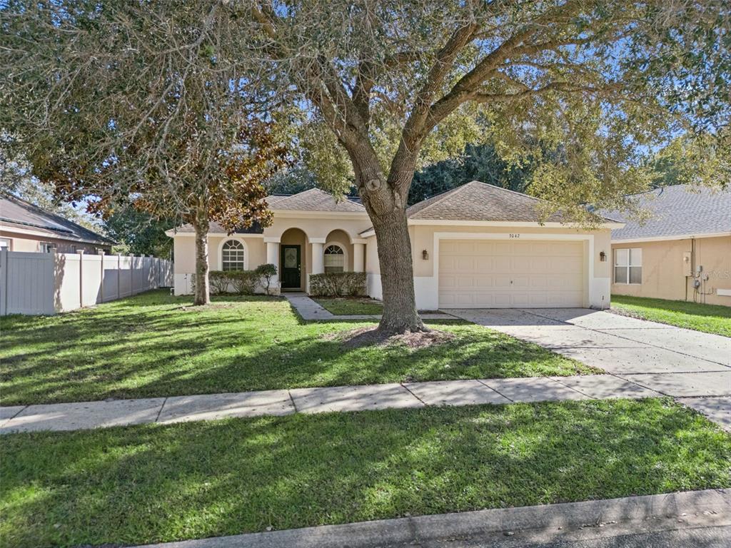 A single-story suburban house with a beige stucco exterior, a two-car garage, and large trees — 3042 WILD PEPPER AVENUE, DELTONA, FL