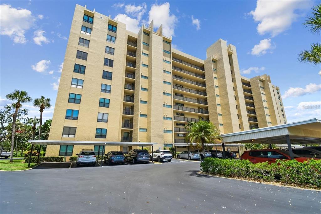 A tall, tan multi-story apartment building with covered parking spaces in the foreground — 2621 COVE CAY DRIVE UNIT 703, CLEARWATER, FL