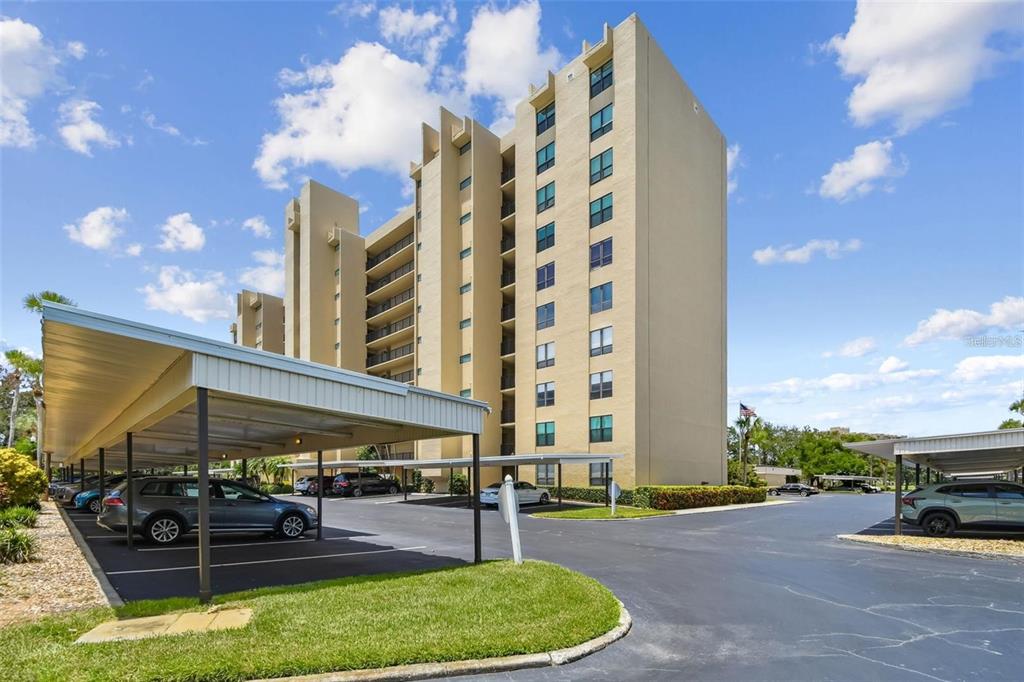 A multi-story tan apartment building with covered parking spaces in the foreground — 2621 COVE CAY DRIVE UNIT 703, CLEARWATER, FL