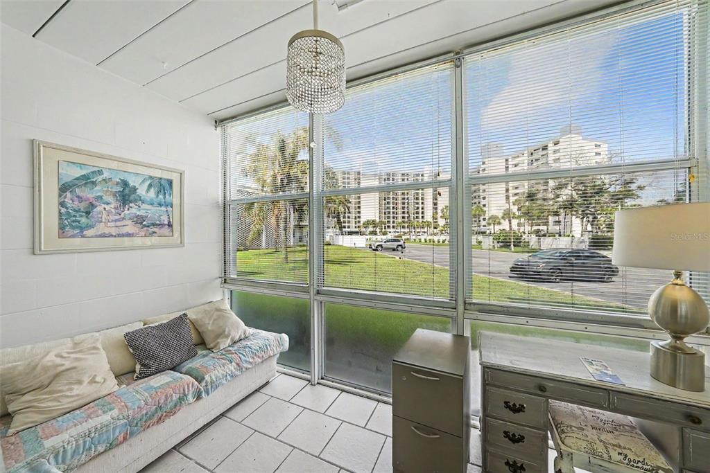 A sunroom features tile floors, a sofa, a desk, and large windows overlooking a parking lot — 7050 SUNSET WAY UNIT 11, ST PETE BEACH, FL
