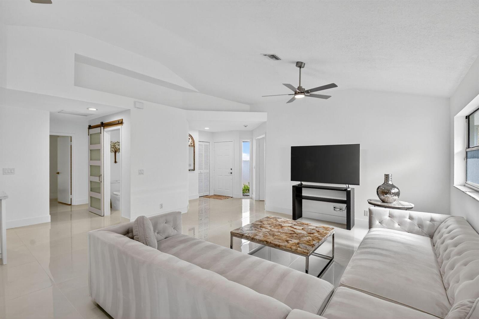 A bright living room features white walls, tile flooring, a sectional sofa, and a television — 2218 NW 52nd Street, Boca Raton FL 33496, BOCA RATON, FL