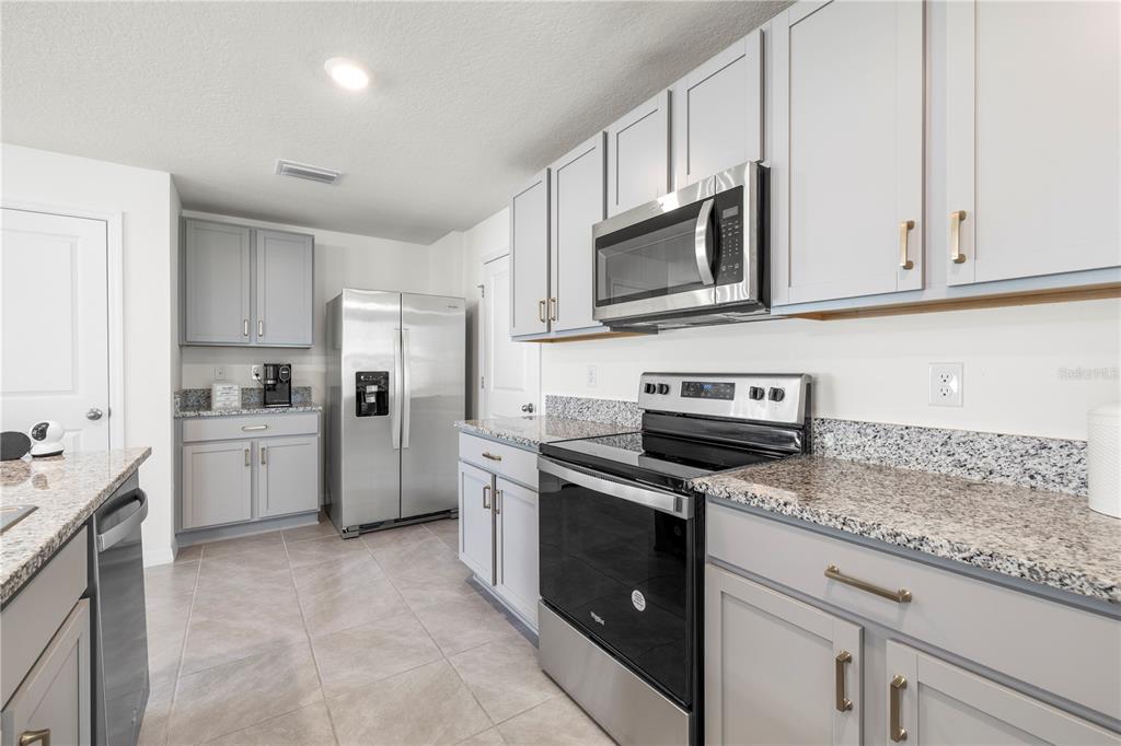 A kitchen features gray cabinets, granite countertops, stainless steel appliances, and light-colored tile flooring — 37247 KINGS CROWN DRIVE, ZEPHYRHILLS, FL