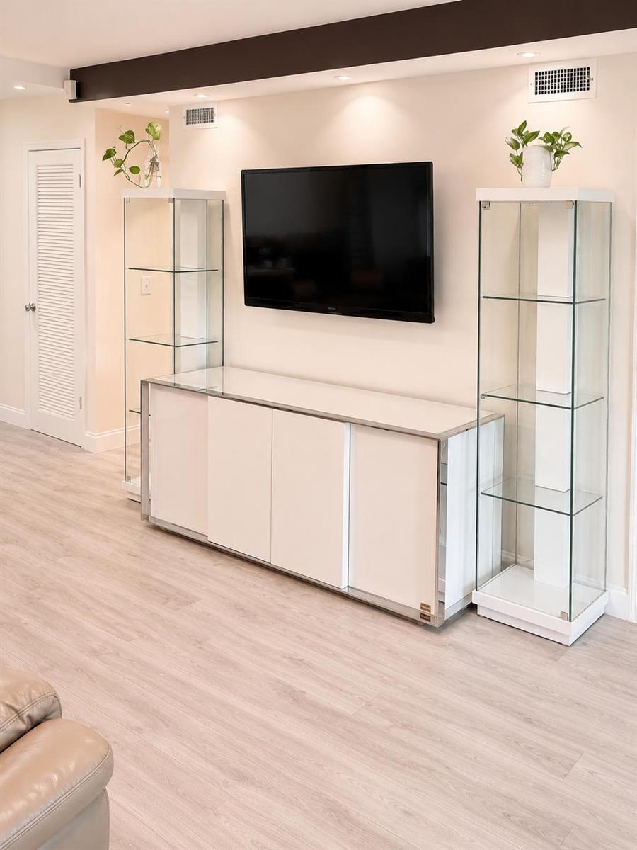 A living room features a wall-mounted television, a white media console, and two glass cabinets — 9880 Marina Boulevard # 1520, Boca Raton FL 33428, BOCA RATON, FL