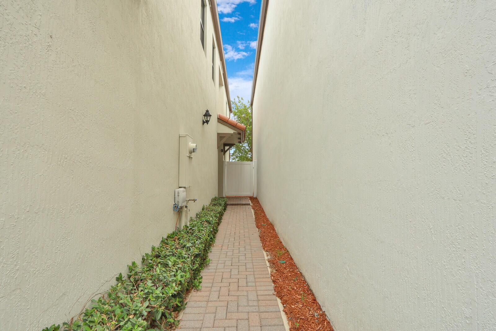 A narrow brick walkway leads between two beige stucco walls toward a white gate — 4008 Sequoia Place, Riviera Beach FL 33410, RIVIERA BEACH, FL