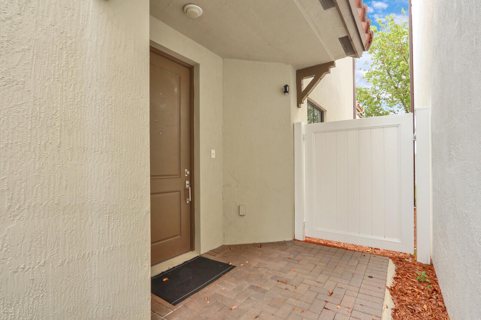 A brown front door opens onto a brick patio enclosed by a white vinyl fence — 4008 Sequoia Place, Riviera Beach FL 33410, RIVIERA BEACH, FL