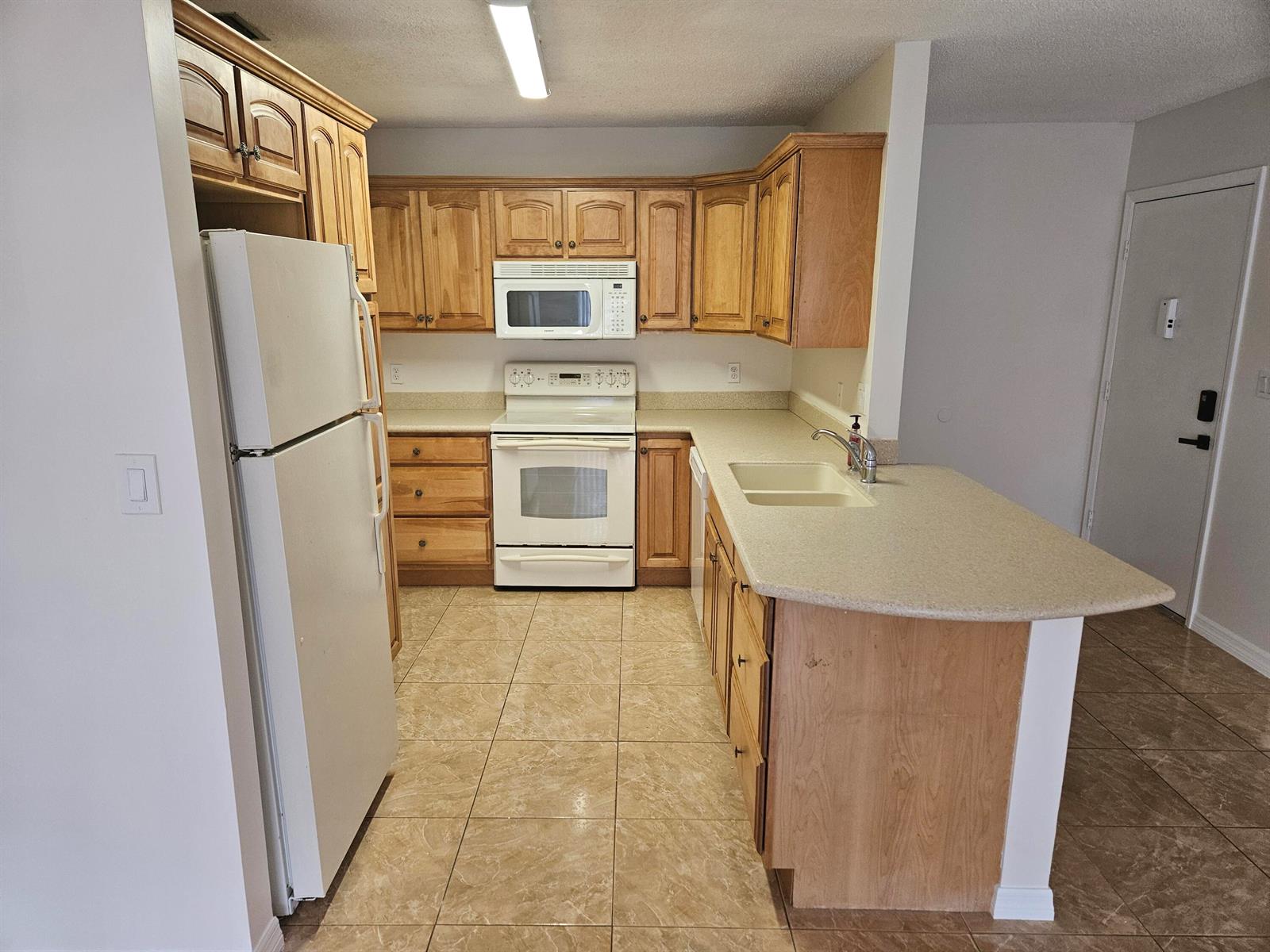 A kitchen features light wood cabinets, white appliances, beige countertops, and tiled flooring — 2050 Oleander Boulevard # 108, Fort Pierce FL 34950, FORT PIERCE, FL