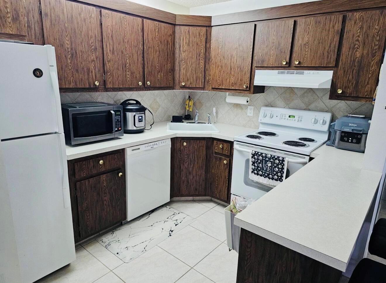 A kitchen features wood cabinets, white appliances, a corner sink, and light-colored tile flooring — 443 Pine Glen Lane # 2, FL 33463, FL