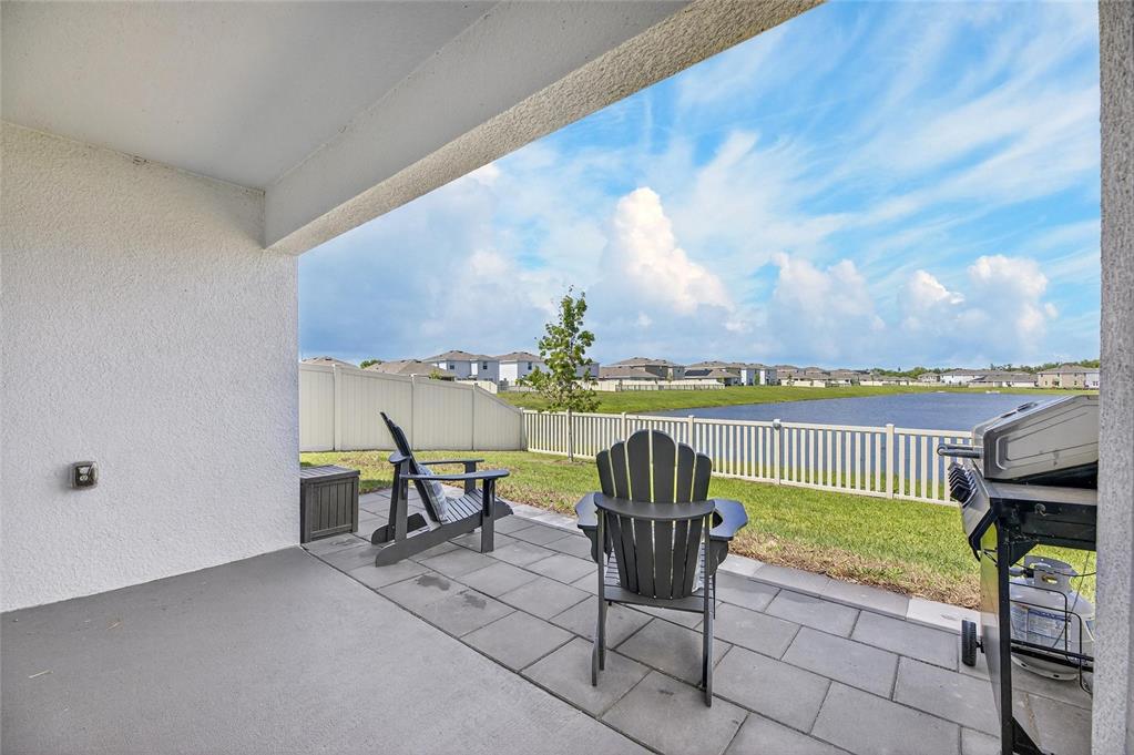 A covered patio features gray pavers, two Adirondack chairs, and a grill overlooking a lake — 3345 BLUE ESTUARY ROAD, WIMAUMA, FL