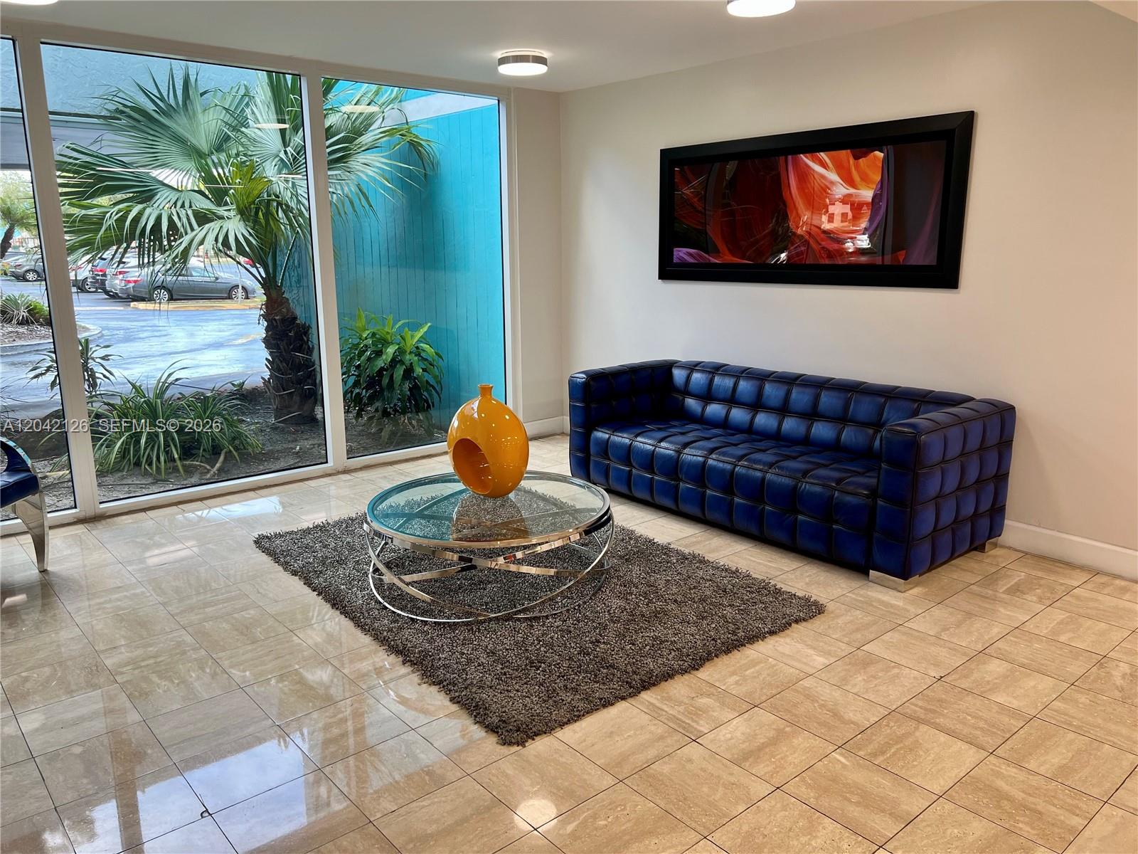 A modern lobby features a blue tufted sofa, glass coffee table, and floor-to-ceiling windows — 919 Hillcrest Dr # 102, Hollywood FL 33021, HOLLYWOOD, FL