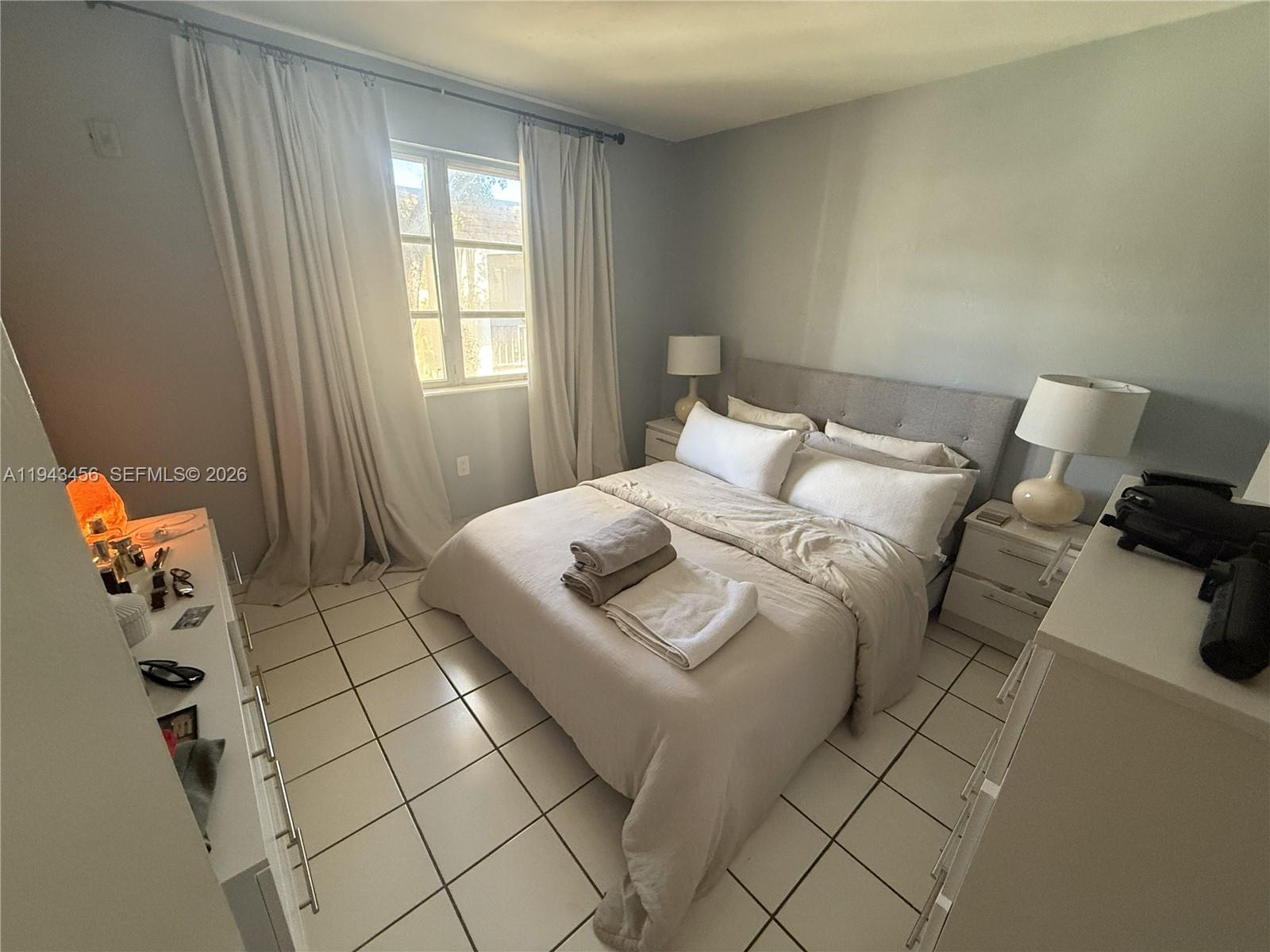 A bedroom features a bed with neutral bedding, tile flooring, and a window with curtains — 7823 Miller Dr # C207, Miami FL 33155, MIAMI, FL
