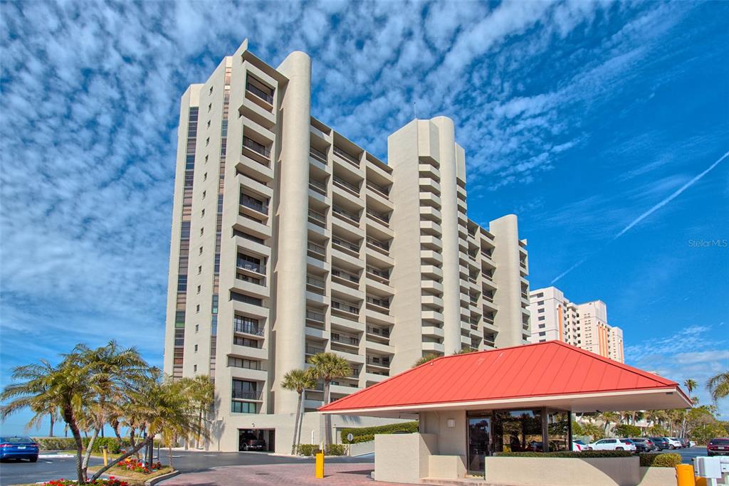 A tall, multi-story beige apartment building stands behind a small guardhouse with a red roof — 1290 GULF BOULEVARD UNIT 903, CLEARWATER BEACH, FL