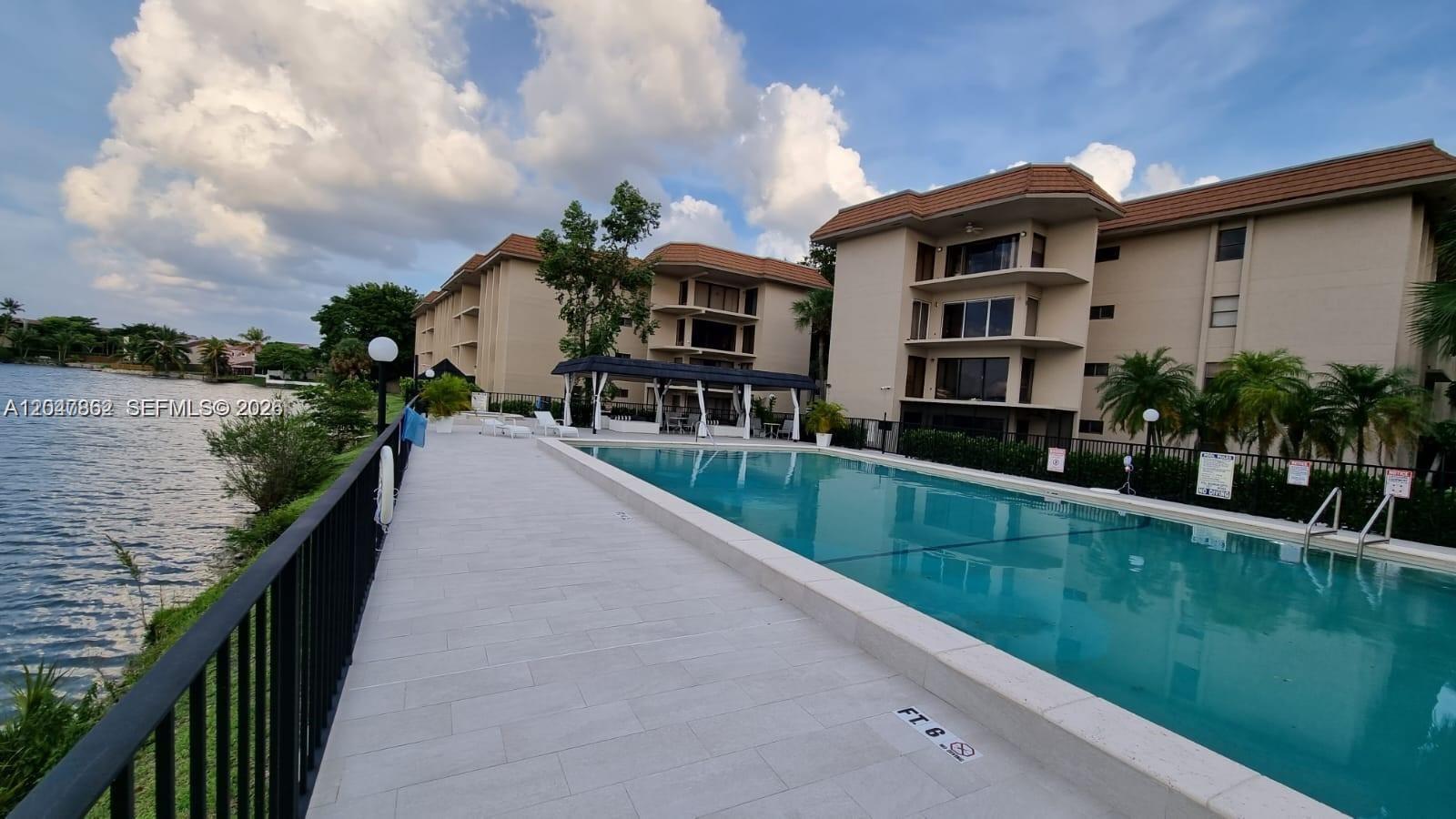 An outdoor swimming pool sits beside a multi-story apartment building and a lake — 461 Ives Dairy Rd # 2, Miami FL 33179, MIAMI, FL