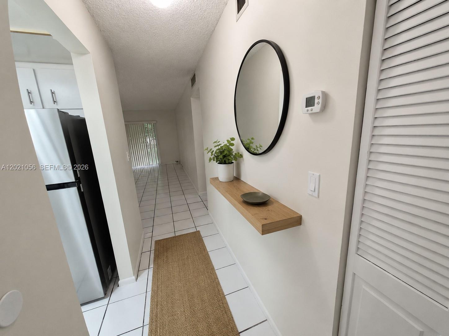 A hallway features white tile floors, a floating wooden shelf, a round mirror, and a rug — 7830 Camino Real # 110, Miami FL 33143, MIAMI, FL