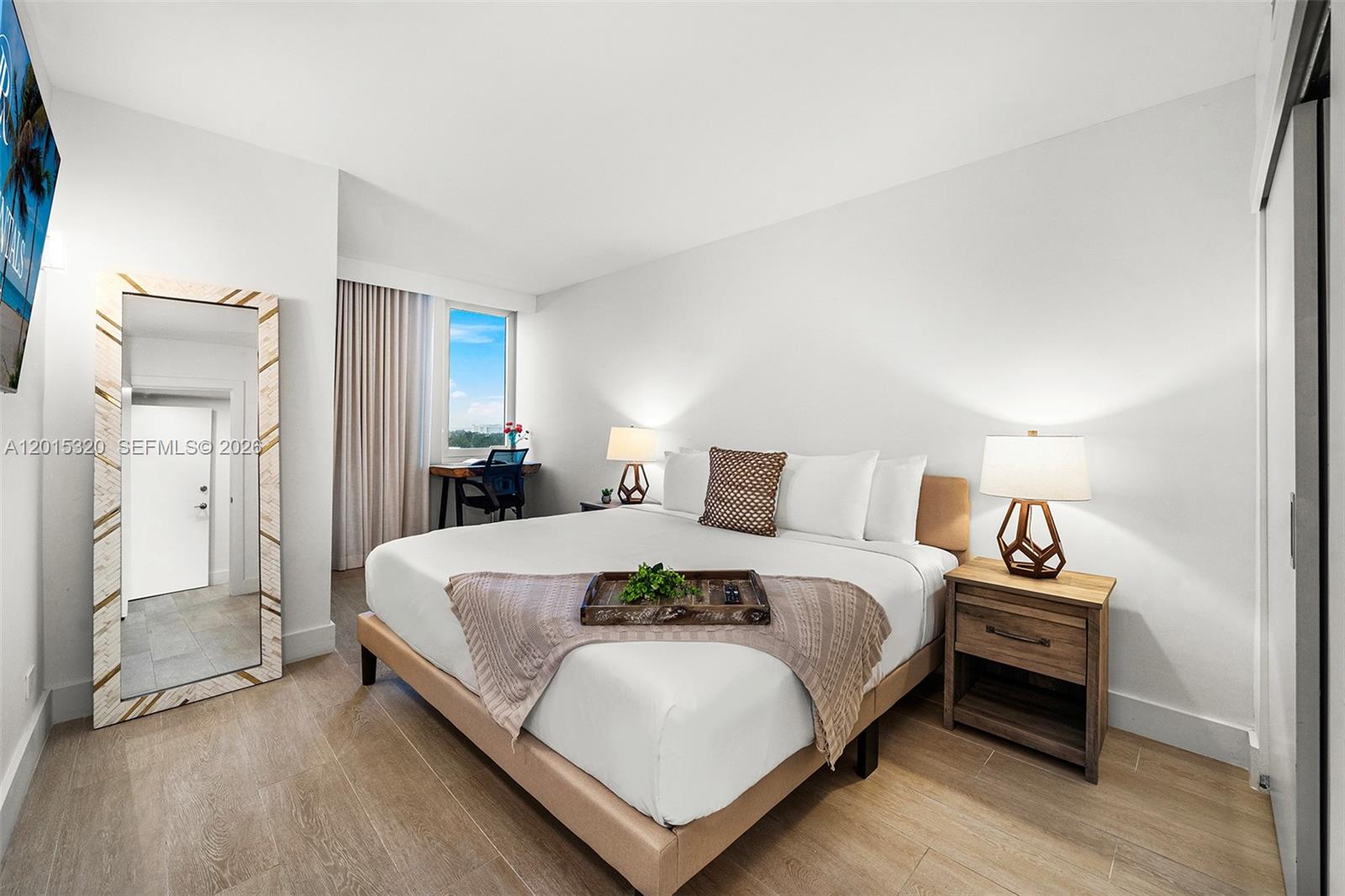 A bedroom features a king bed, wooden nightstand, floor mirror, and a small desk by the window — 2301 Collins Ave # 1129, Miami Beach FL 33139, MIAMI BEACH, FL