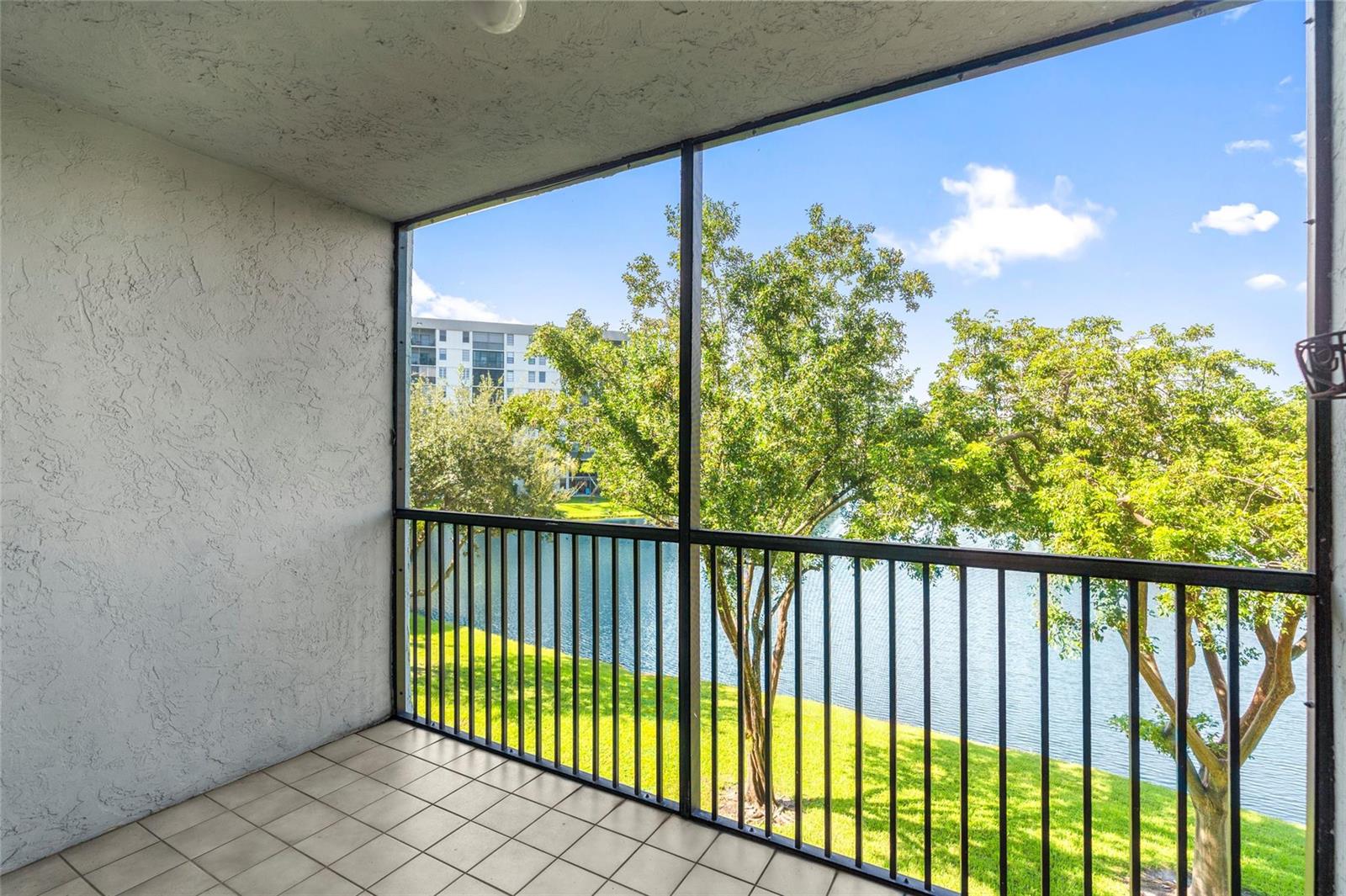 A screened-in balcony with tiled flooring overlooks a grassy lawn, a lake, and trees — 2226 N Cypress Bend Drive # 308, Pompano Beach FL 33069, POMPANO BEACH, FL