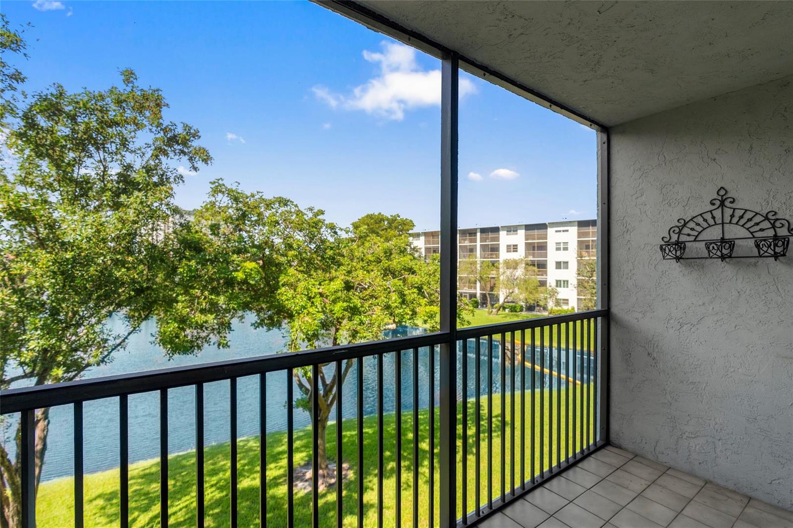 A screened-in balcony with tile flooring overlooks a lake, green lawn, and apartment buildings — 2226 N Cypress Bend Drive # 308, Pompano Beach FL 33069, POMPANO BEACH, FL