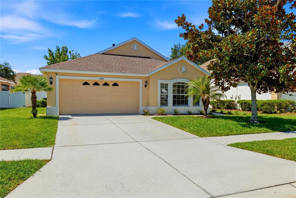 A single-story tan house with a two-car garage, a concrete driveway, and a front lawn — 19406 SUNSET BAY DRIVE, LAND O LAKES, FL