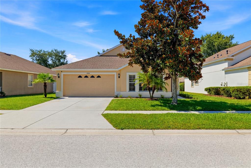 A single-story house with a tan exterior, a two-car garage, and a large front tree — 19406 SUNSET BAY DRIVE, LAND O LAKES, FL