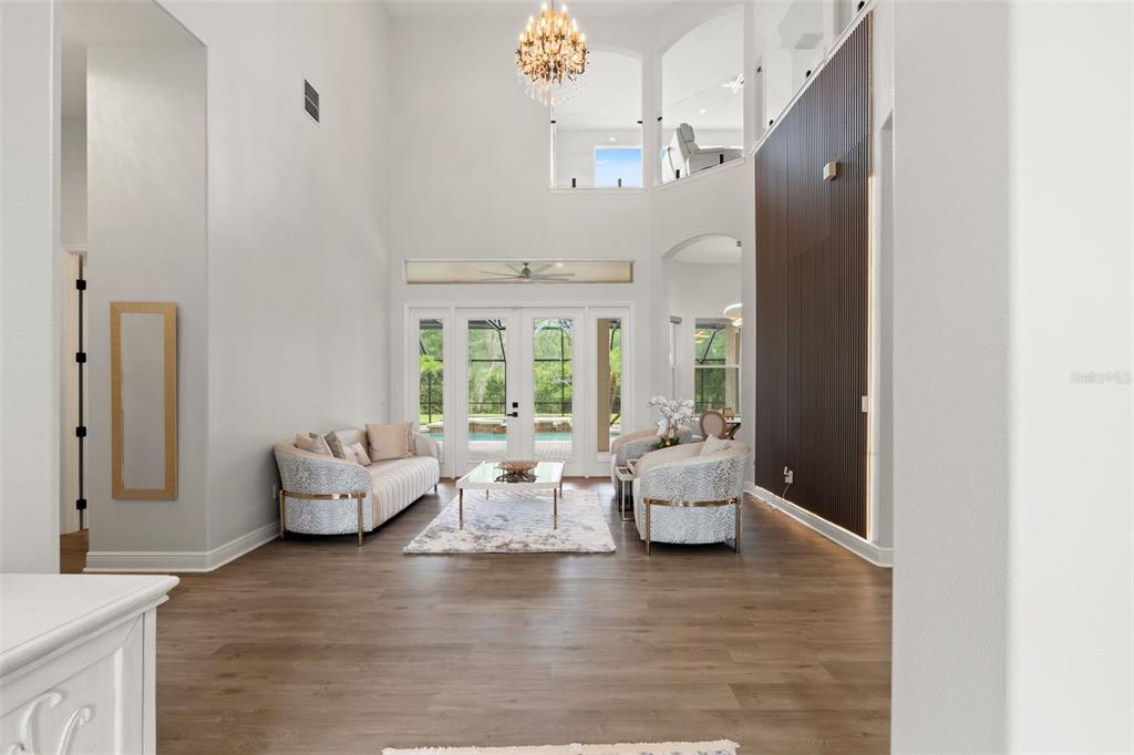 A bright, high-ceilinged living room features wood-look flooring, a large chandelier, and a slatted accent wall — 8412 DUNHAM STATION DRIVE, TAMPA, FL
