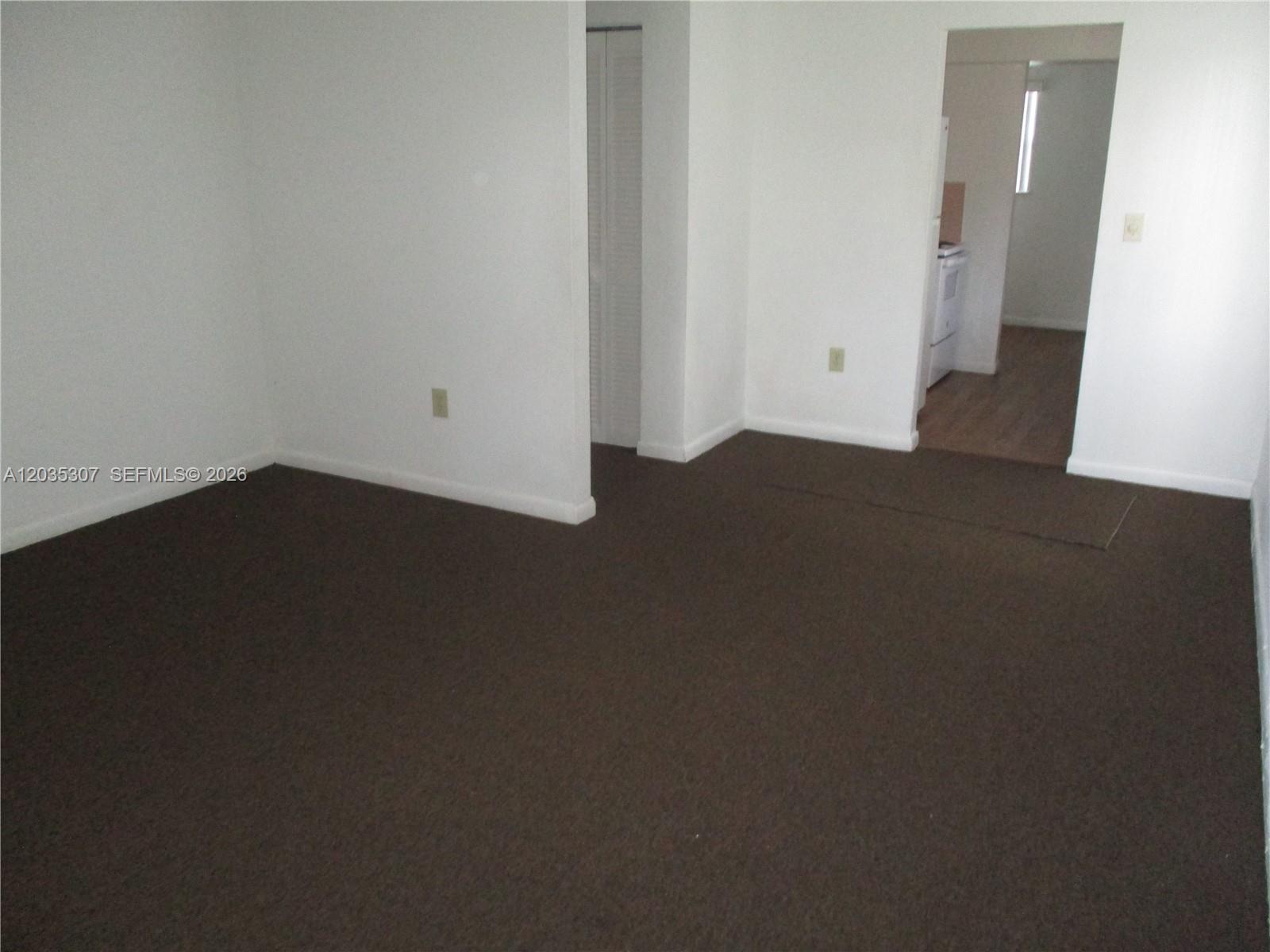 An empty room with dark brown carpeting and white walls leads into a kitchen area — 10622 SW 174th Ter # 10622, Miami FL 33157, MIAMI, FL