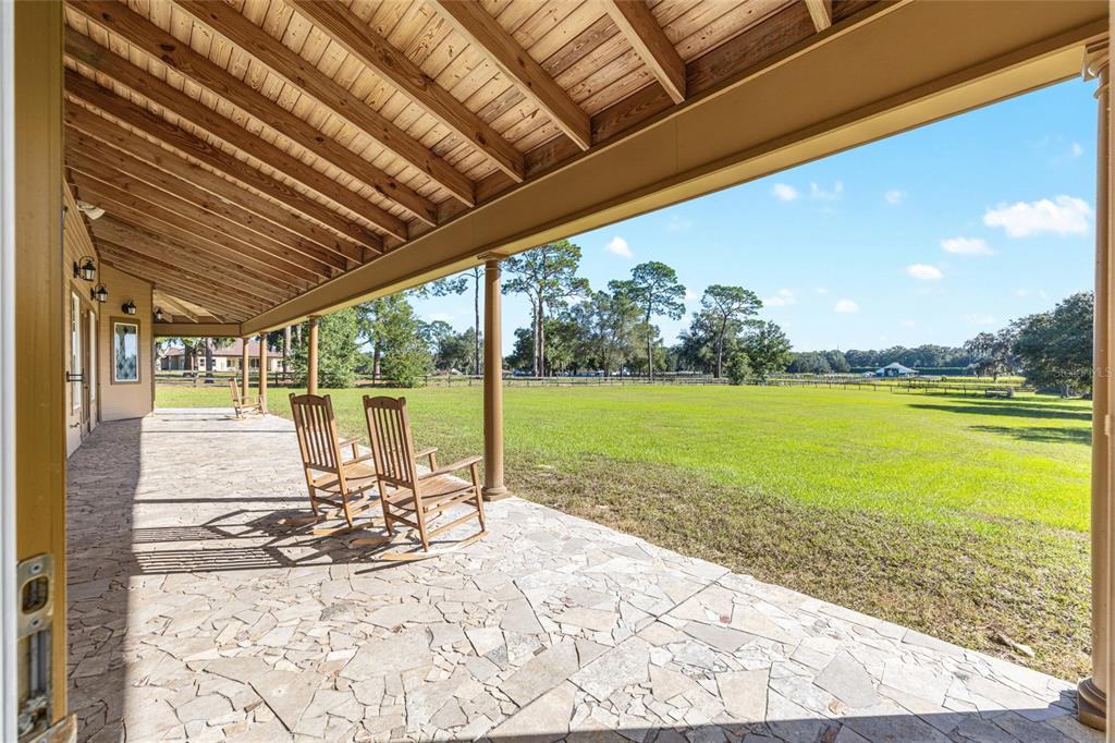 A covered stone patio with rocking chairs overlooks a large grassy field and trees — 997 NW 63RD COURT, OCALA, FL