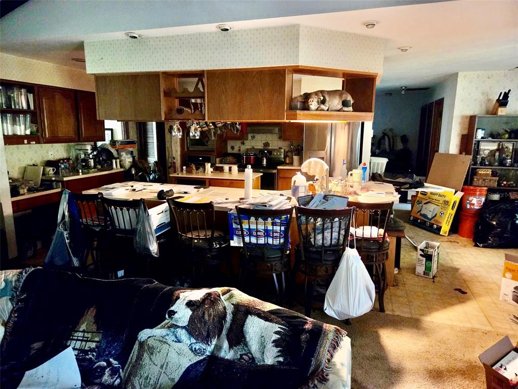 A cluttered kitchen features wood cabinets, a central island with bar seating, and patterned wallpaper — 741 BRECHNER TERRACE, DELTONA, FL