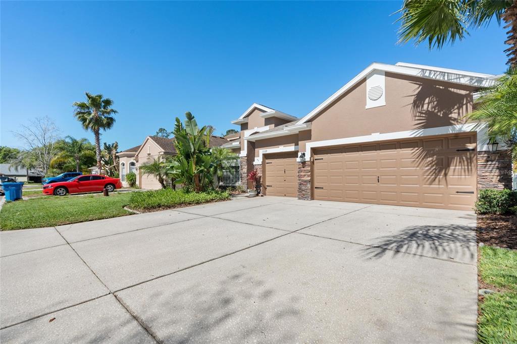 A tan suburban house with a three-car garage and a concrete driveway under a blue sky — 11129 OYSTER BAY CIRCLE, NEW PORT RICHEY, FL