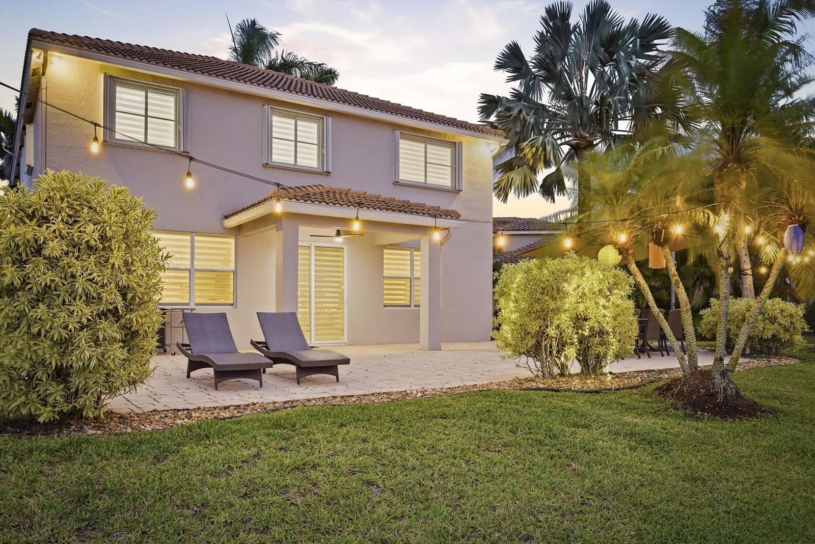 A two-story house exterior with a paved patio, lounge chairs, and string lights at dusk — 1913 Silverbell Terrace, Weston FL 33327, WESTON, FL