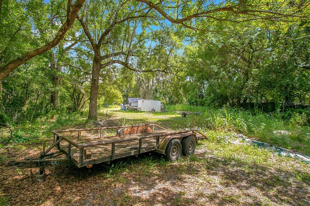 A flatbed trailer sits on a grassy lot surrounded by trees and overgrown vegetation — 8816 TREVARTHON ROAD, ORLANDO, FL