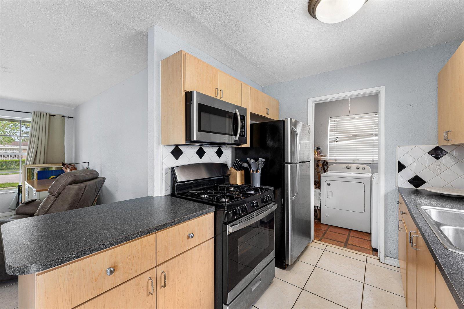 A kitchen features light wood cabinets, black appliances, a dark countertop, and a laundry room beyond — 5543 SW 7th Street, Margate FL 33068, MARGATE, FL