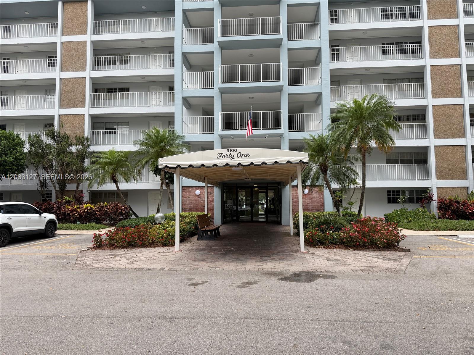 The exterior of a multi-story apartment building features a covered entrance with a bench — 3100 N Course Ln # 208, Pompano Beach FL 33069, POMPANO BEACH, FL