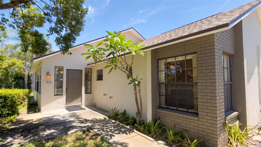 The exterior of a single-story house features a tan stucco facade, brick accents, and landscaping — 7636 HAIG COURT, NEW PORT RICHEY, FL
