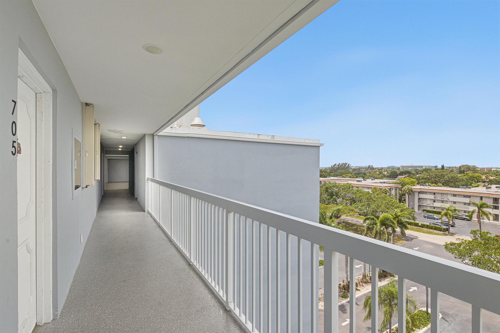 An outdoor apartment building walkway with a white railing overlooks a parking lot and trees — 5700 NW 2nd Avenue # 705, Boca Raton FL 33487, BOCA RATON, FL