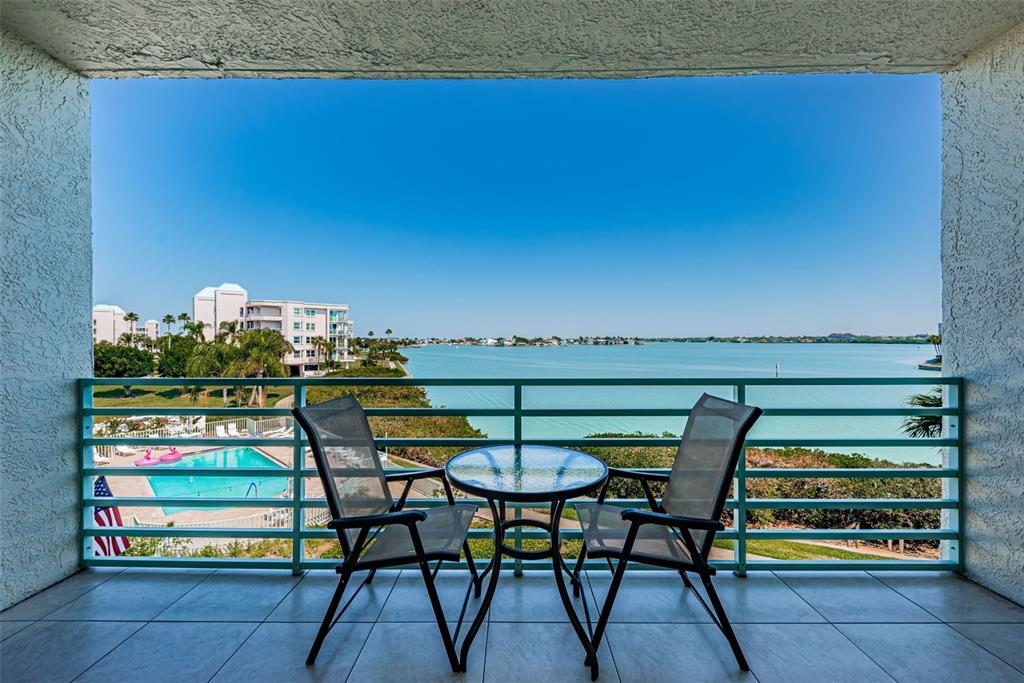 A balcony with a glass-top table and two chairs overlooks a swimming pool and bay — 7979 SAILBOAT KEY BOULEVARD S UNIT 205, SOUTH PASADENA, FL
