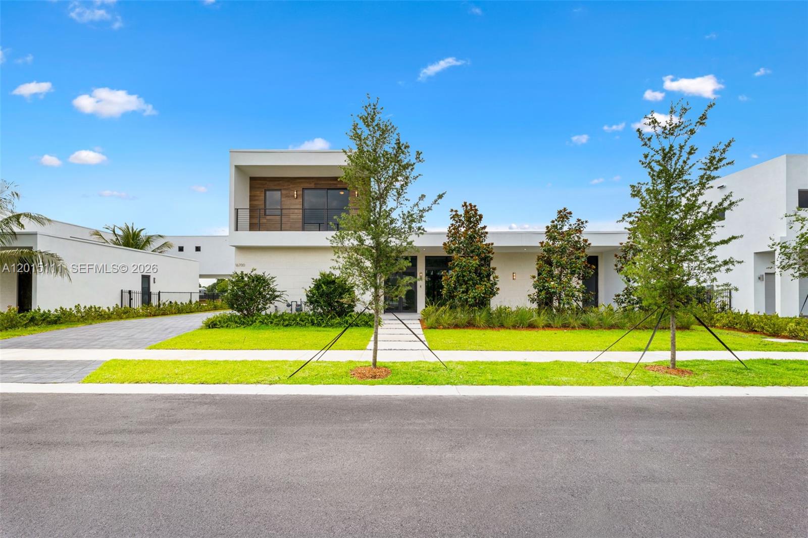 A modern white house with a wood-accented balcony stands behind a manicured lawn and trees — 16700 N Botaniko Dr N, Weston FL 33326, WESTON, FL