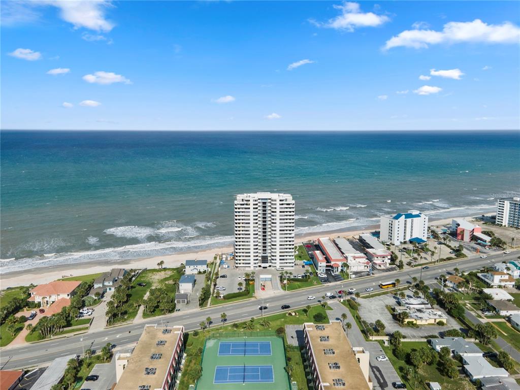 An aerial view shows a high-rise condominium building on a beach near tennis courts — 1420 N ATLANTIC AVENUE UNIT 703, DAYTONA BEACH, FL