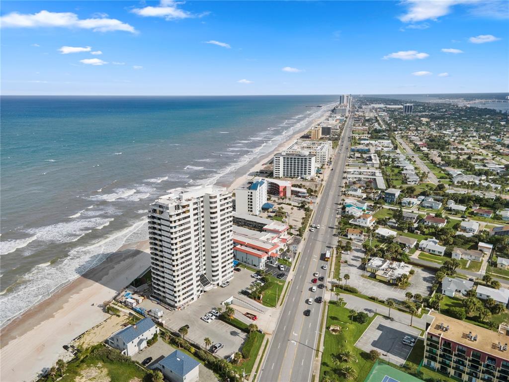 An aerial view shows a high-rise building along a beach next to a coastal road — 1420 N ATLANTIC AVENUE UNIT 703, DAYTONA BEACH, FL