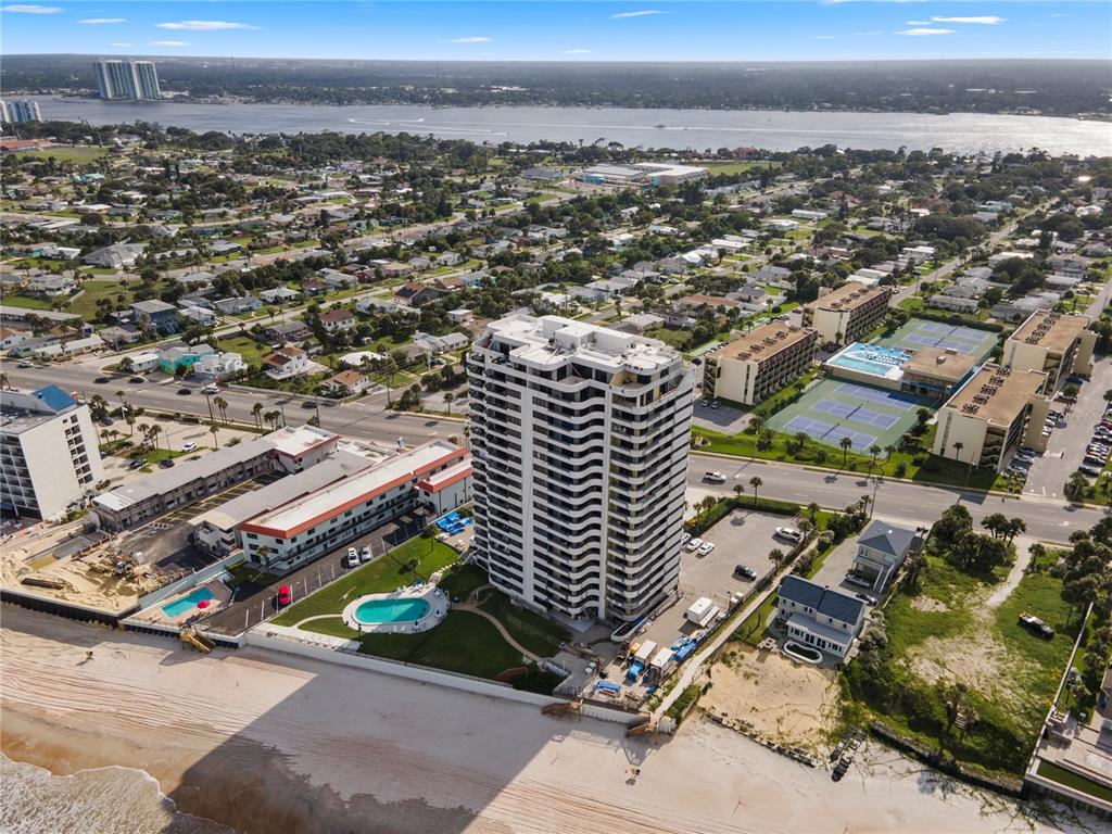 An aerial view shows a tall beachfront condominium tower surrounded by parking, pools, and tennis courts — 1420 N ATLANTIC AVENUE UNIT 703, DAYTONA BEACH, FL