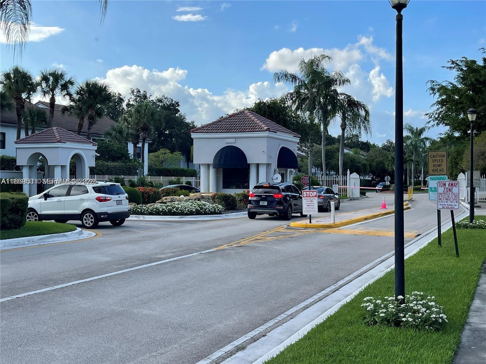 A gated entrance with a guardhouse, palm trees, and cars waiting to enter the community — 5133 Brisata # L, Boynton Beach FL 33437, BOYNTON BEACH, FL