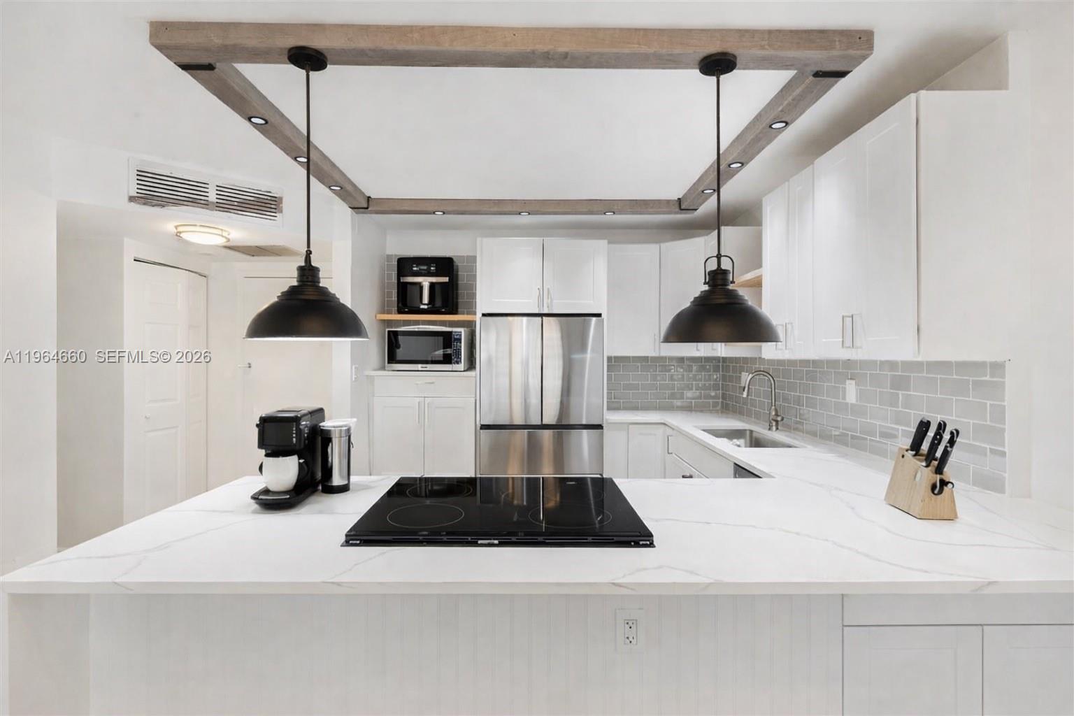 A modern kitchen features white cabinetry, a marble-look island with a cooktop, and wooden ceiling beams — 600 Three Islands Blvd # 1214, Hallandale Beach FL 33009, HALLANDALE BEACH, FL