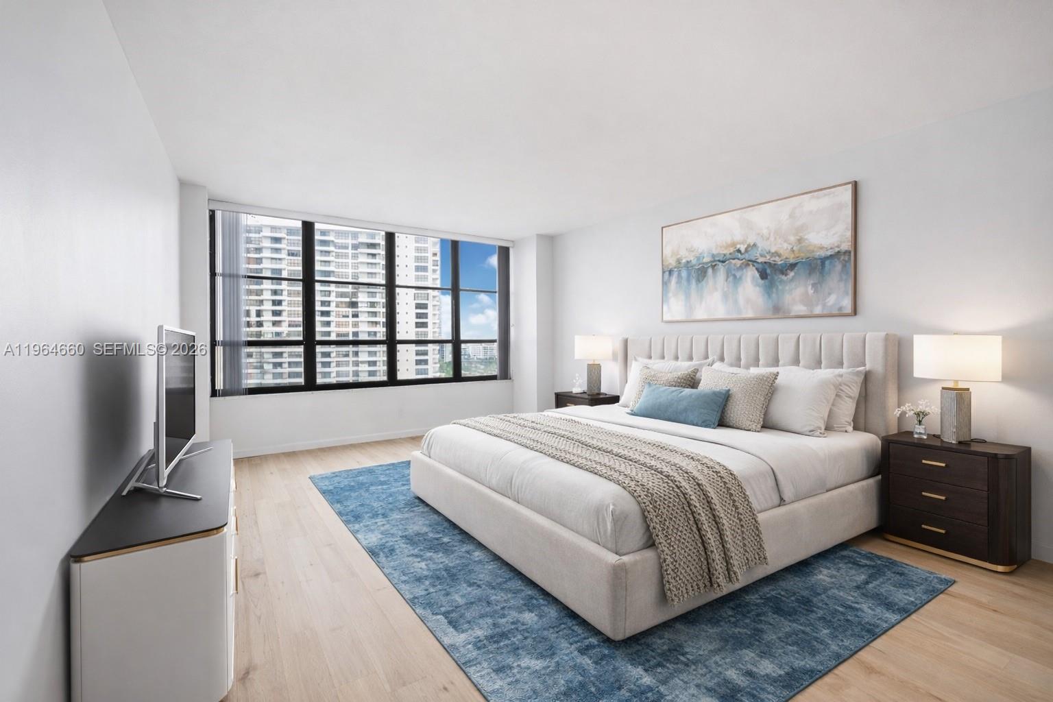 A modern bedroom features a king bed, blue area rug, and large windows with city views — 600 Three Islands Blvd # 1214, Hallandale Beach FL 33009, HALLANDALE BEACH, FL