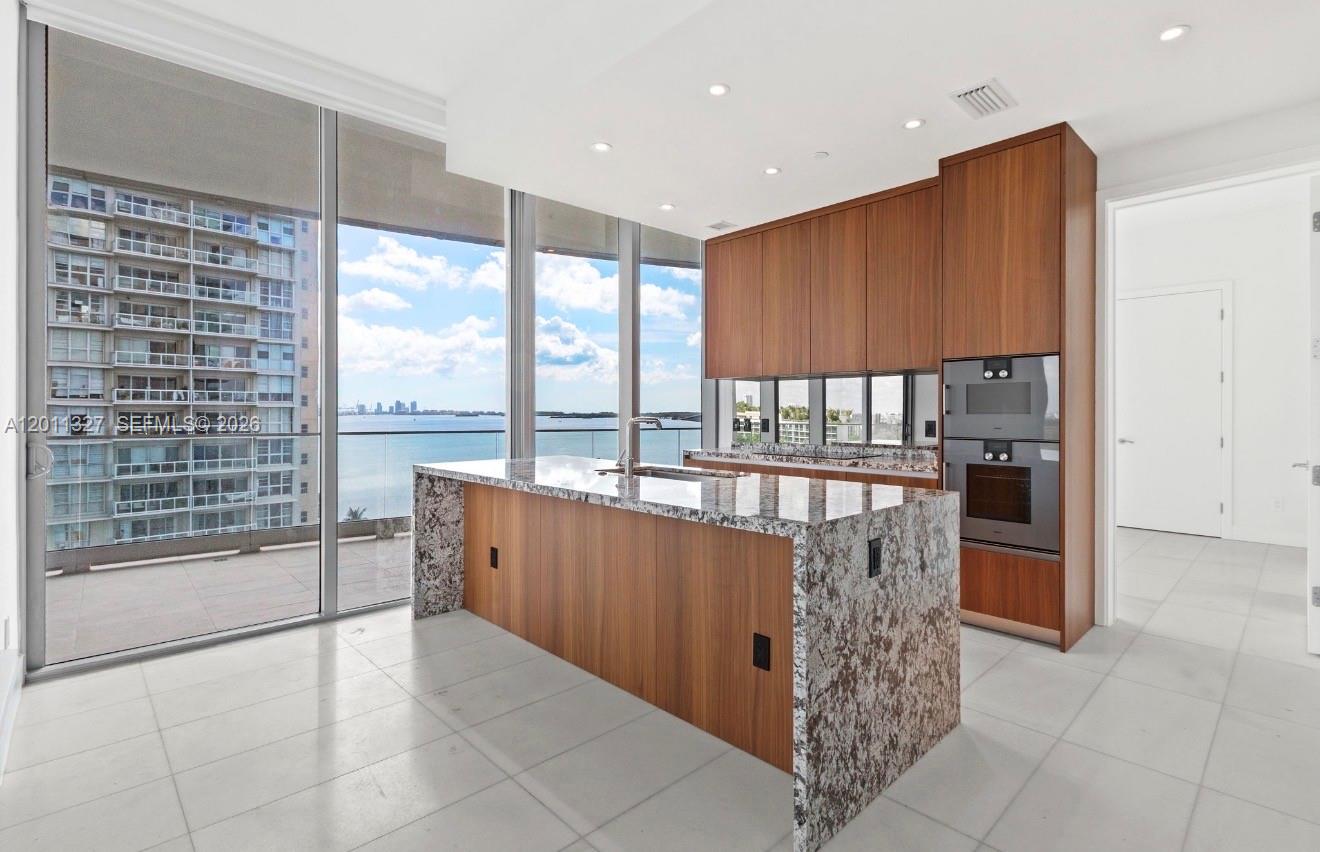 A modern kitchen features wood cabinetry, a granite waterfall island, and floor-to-ceiling windows overlooking water — 175 SE 25th Rd # 704, Miami FL 33129, MIAMI, FL