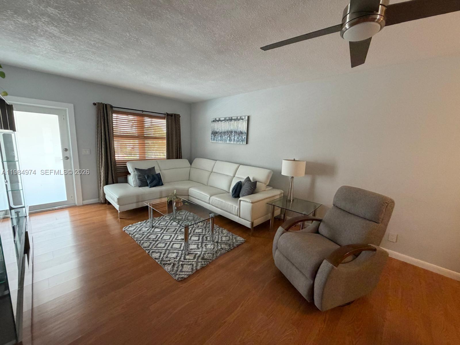 A living room features a white sectional sofa, a gray recliner, and wood-look flooring — 630 Layne Blvd # 215, Hallandale Beach FL 33009, HALLANDALE BEACH, FL