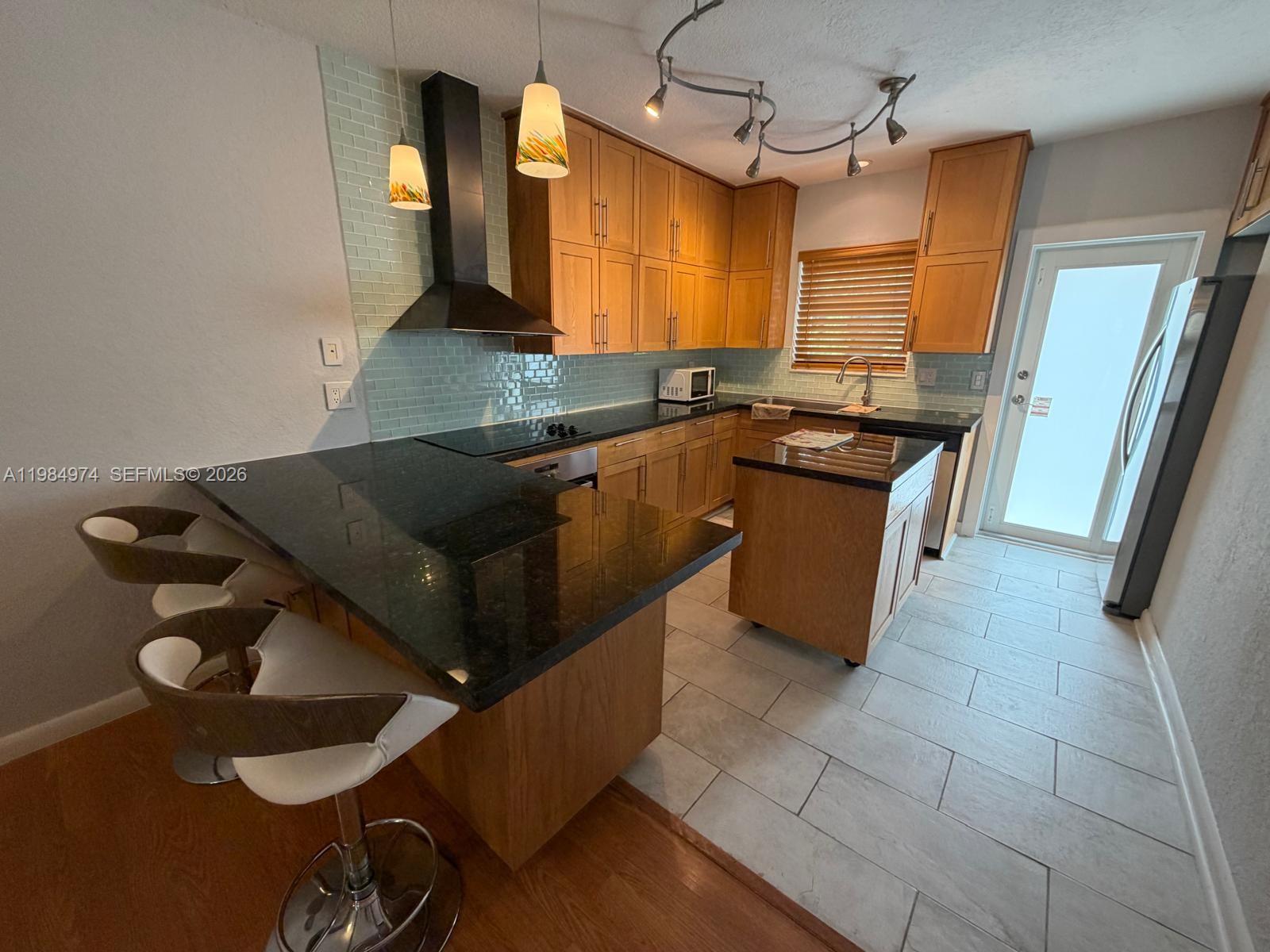 A kitchen features light wood cabinets, black granite countertops, a tile backsplash, and a mobile island — 630 Layne Blvd # 215, Hallandale Beach FL 33009, HALLANDALE BEACH, FL